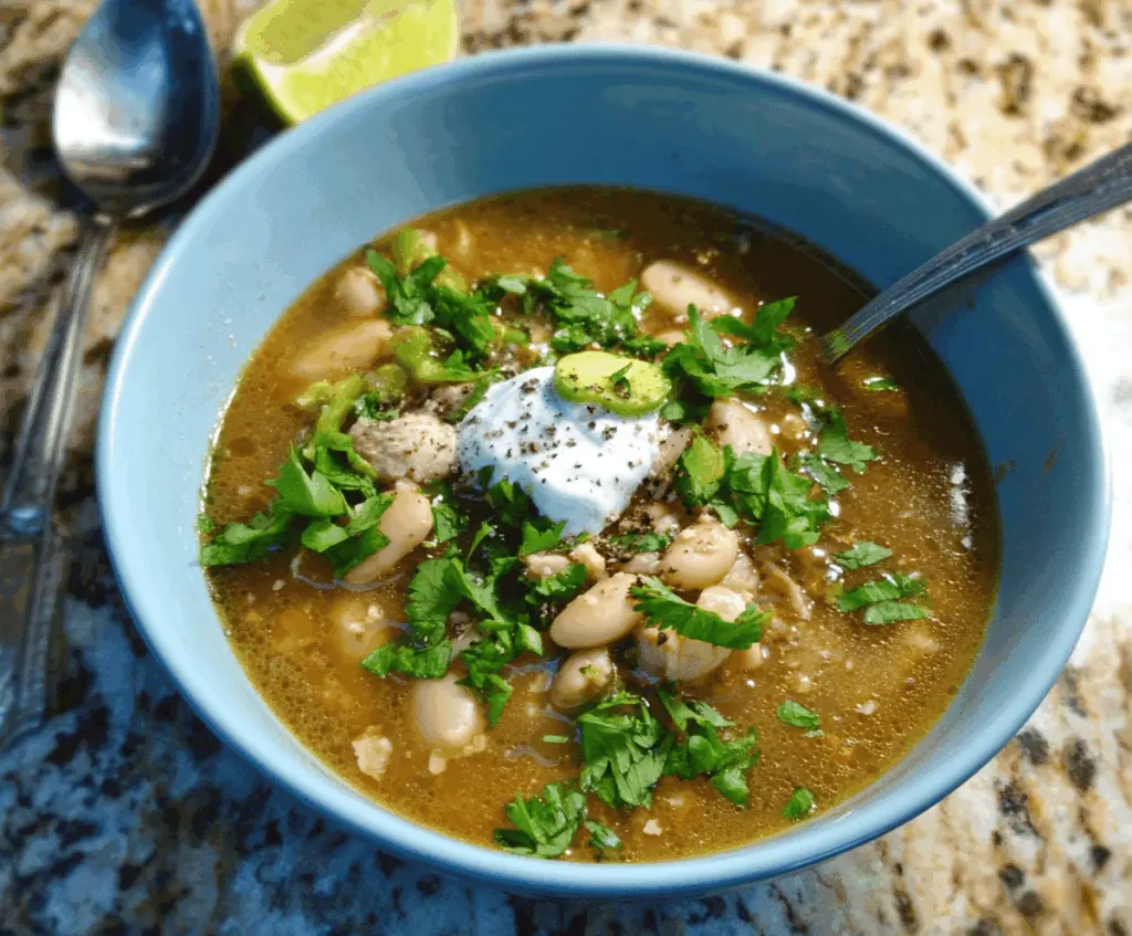 Creamy white chili topped with shredded cheese, fresh cilantro, and diced green chilies in a white bowl on a rustic wooden table.